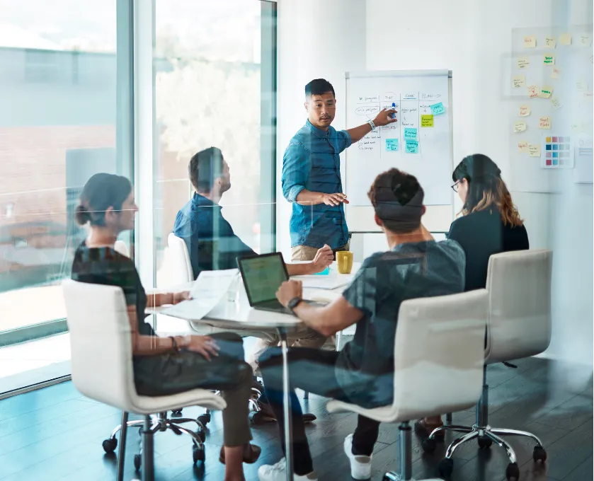 about-1 A man leads a team meeting, presenting ideas on a whiteboard in a modern office setting.