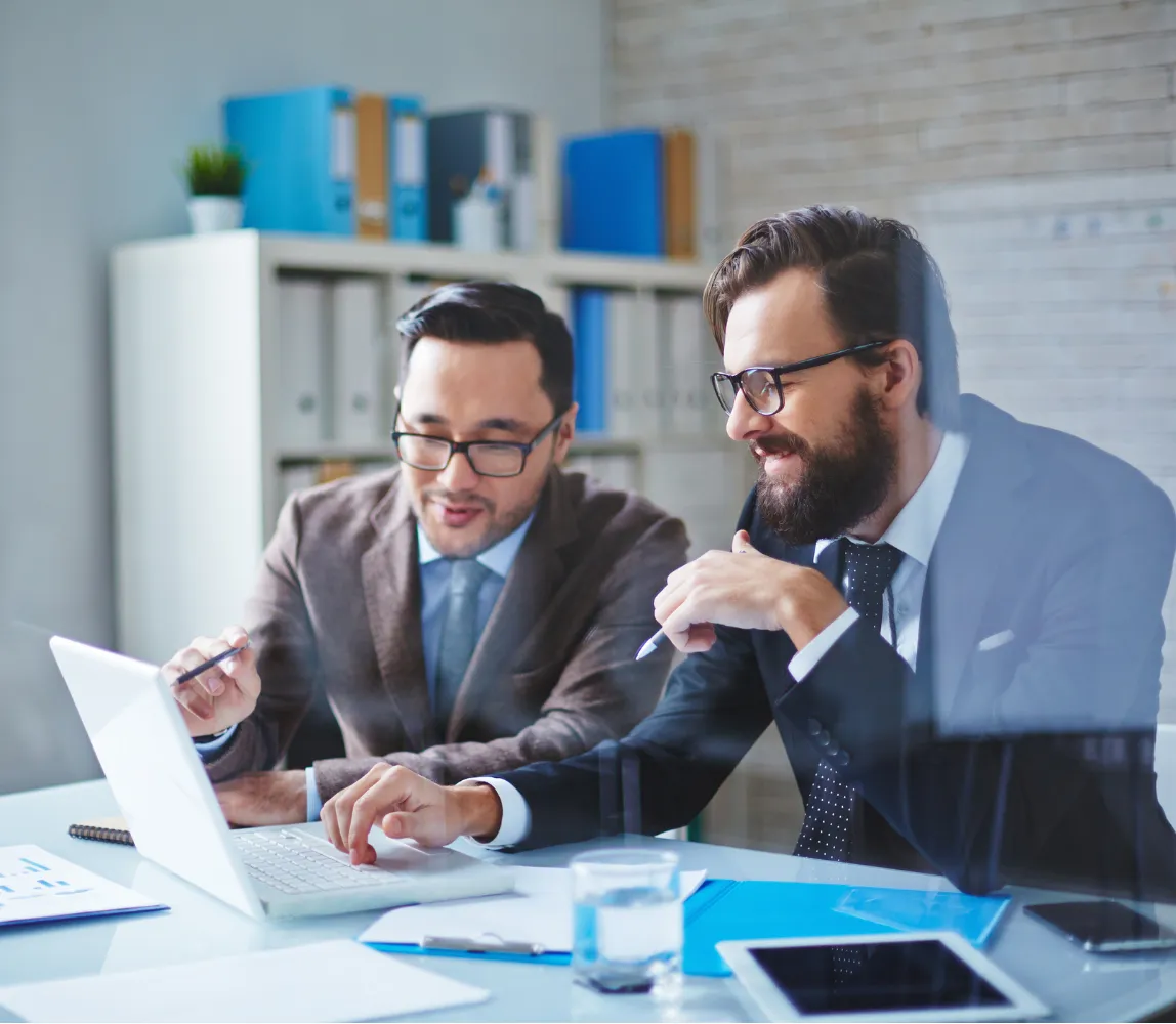 Two businessmen in suits working together at a desk, looking at a laptop screen in an office.