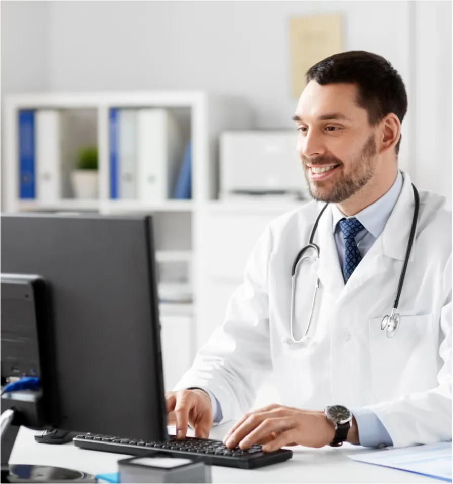 Smiling doctor in a white coat working at a computer in a modern medical office.