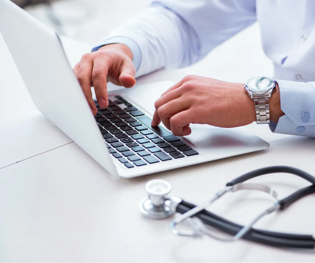 ehr-1 Person typing on a laptop at a desk, with a stethoscope lying nearby.