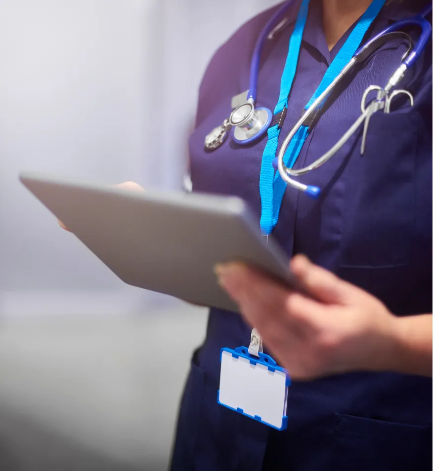 A healthcare professional in scrubs holds a tablet, with a stethoscope and ID badge visible.