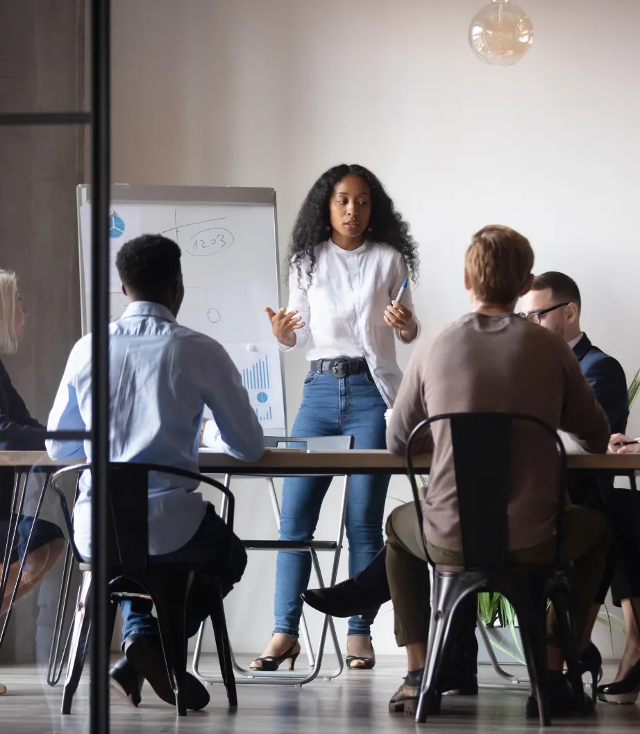 Woman giving a presentation to a group of colleagues in an office meeting room with a whiteboard.