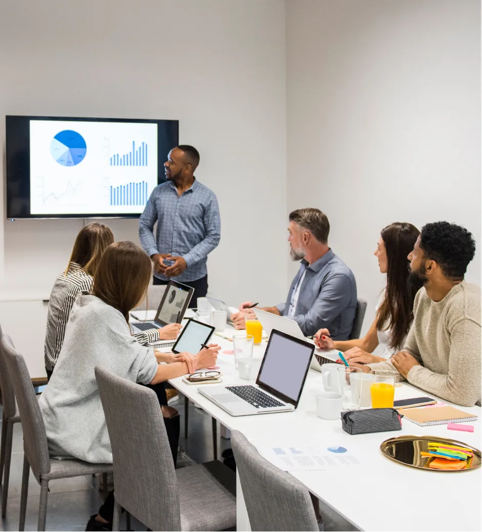 A man presents charts on a screen to five colleagues seated at a conference table with laptops and papers.