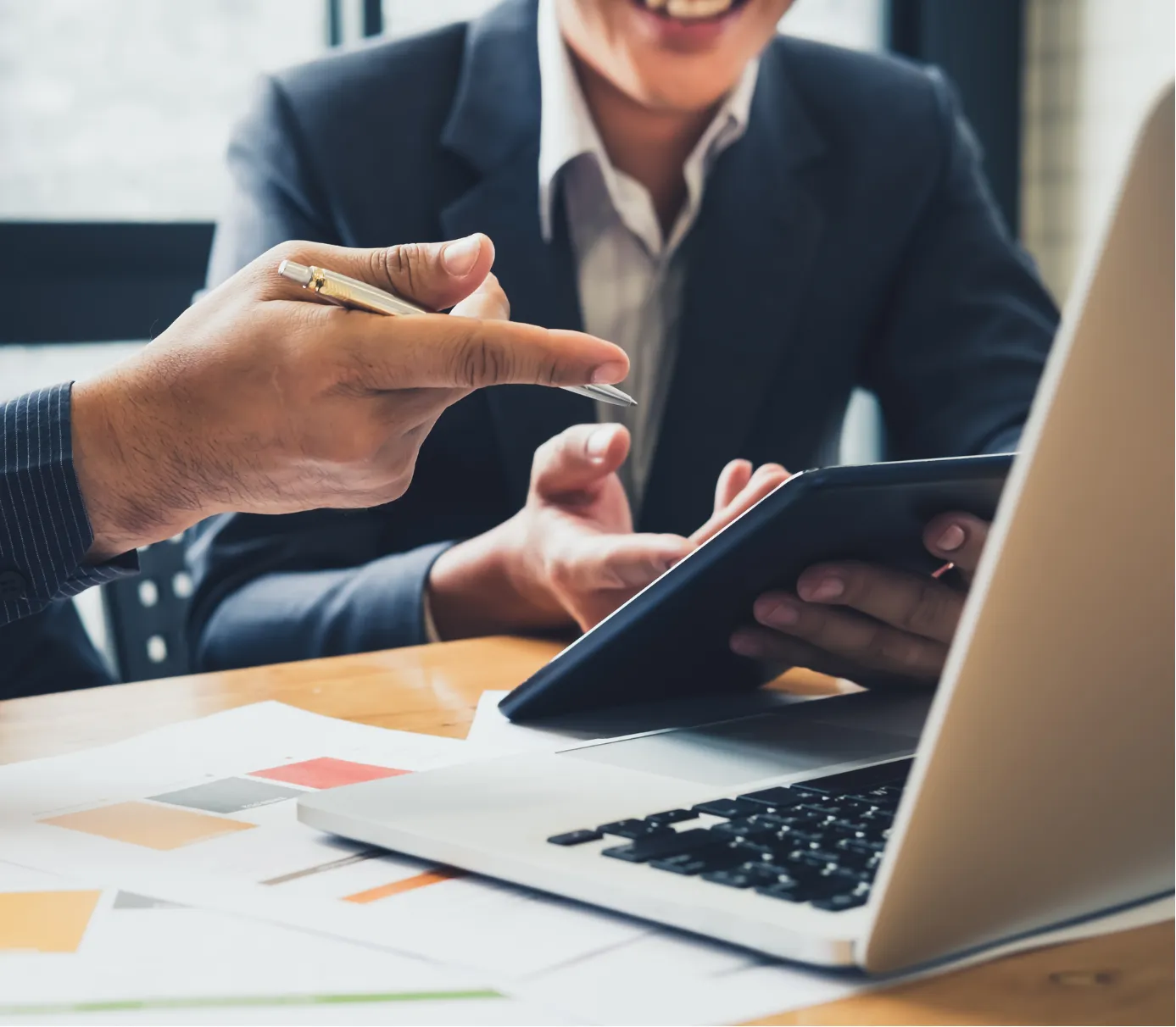 Two people in business suits discuss work at a desk with a laptop, tablet, and documents.