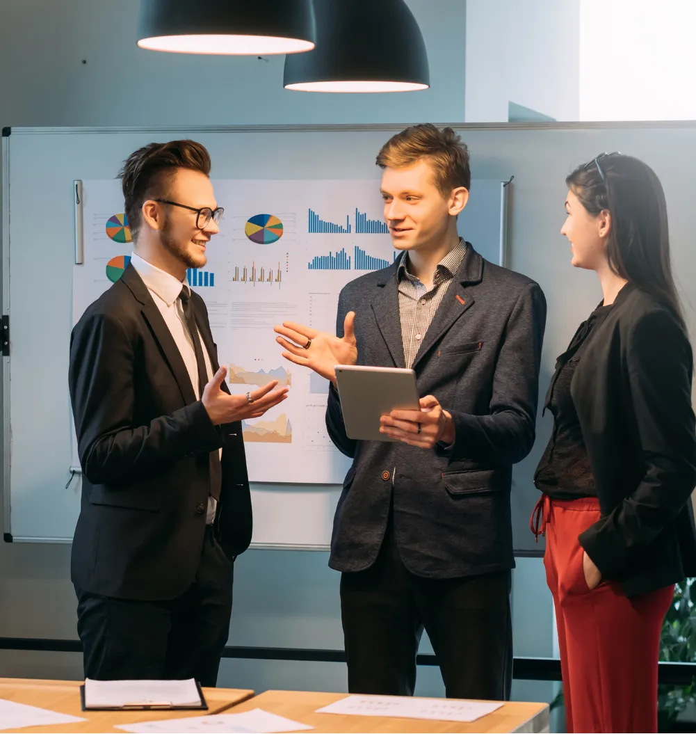 Three colleagues in business attire discuss work in front of a whiteboard with charts and graphs.