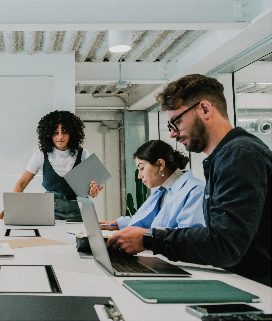 Three people working at a table with laptops in a modern office; one person stands holding a laptop.