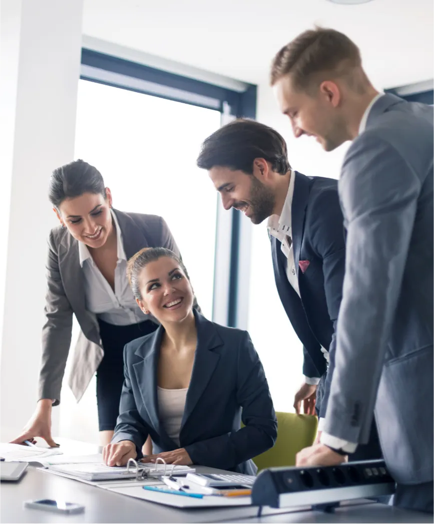 Four business professionals in suits smiling and collaborating around a desk in a bright office.