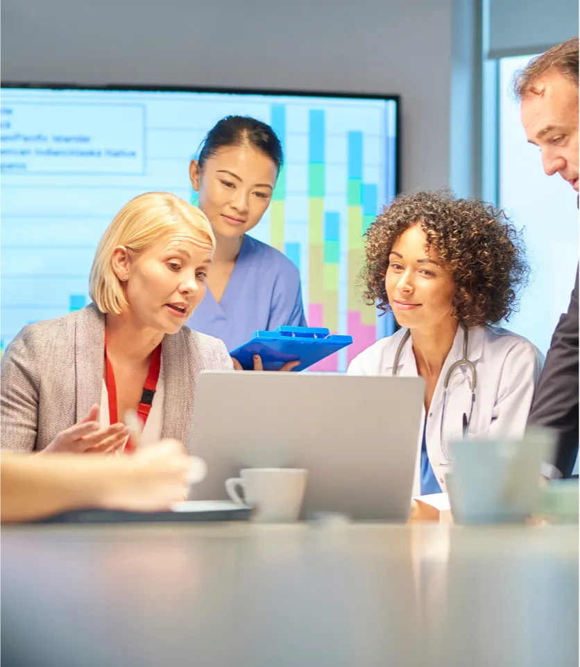 req-2 Four healthcare professionals discuss something while looking at a laptop in a meeting room.