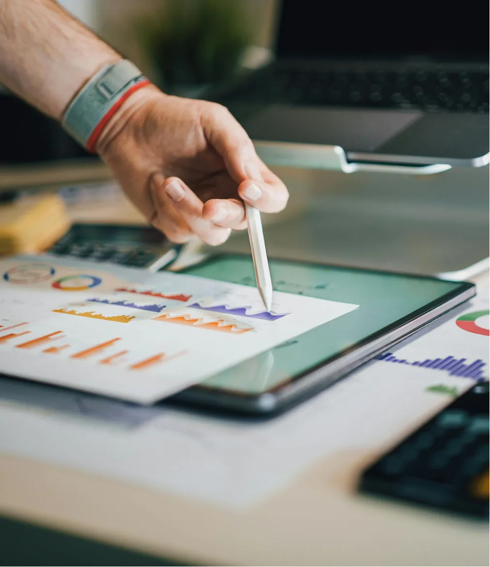 A person points at a printed chart with a pen, using a tablet and analyzing graphs at a desk.