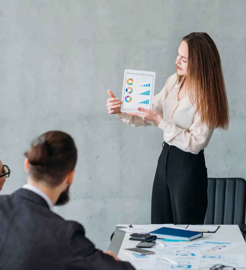 Woman presenting colorful charts on a tablet to colleagues in a modern office meeting.