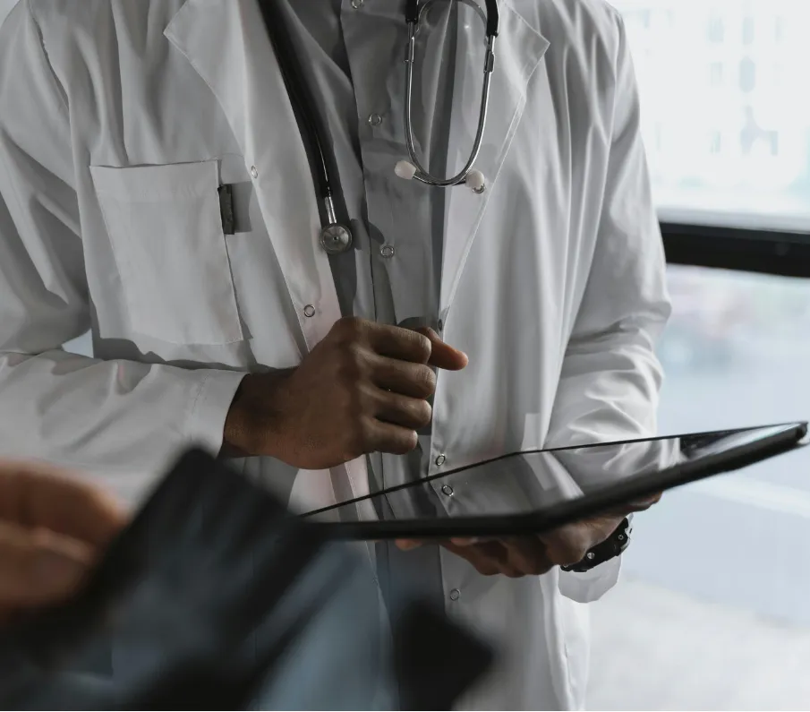 services-1 Doctor in a white coat holding a tablet and wearing a stethoscope, standing near a window.