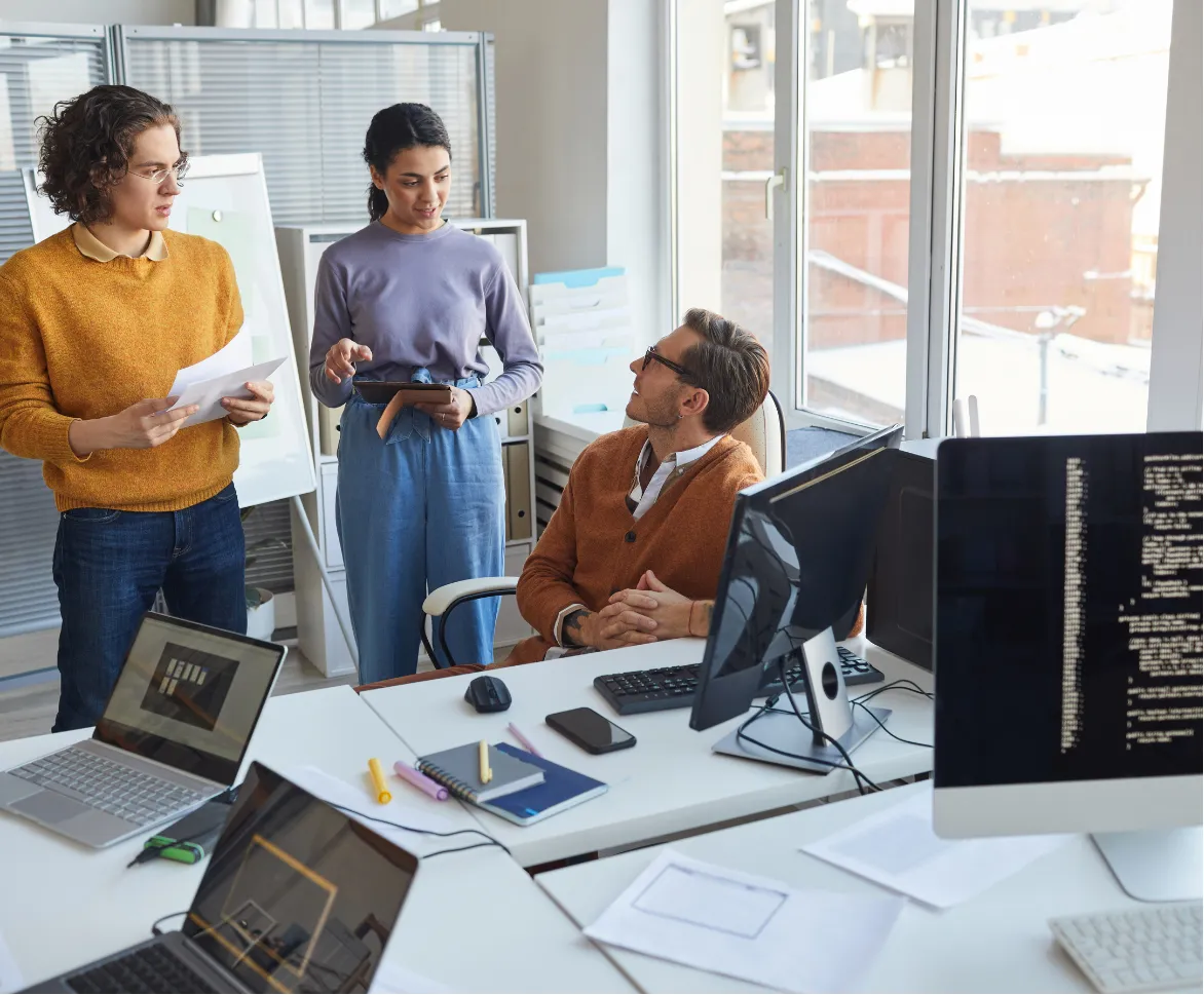Three people have a discussion in a modern office with computers and laptops on the desks.