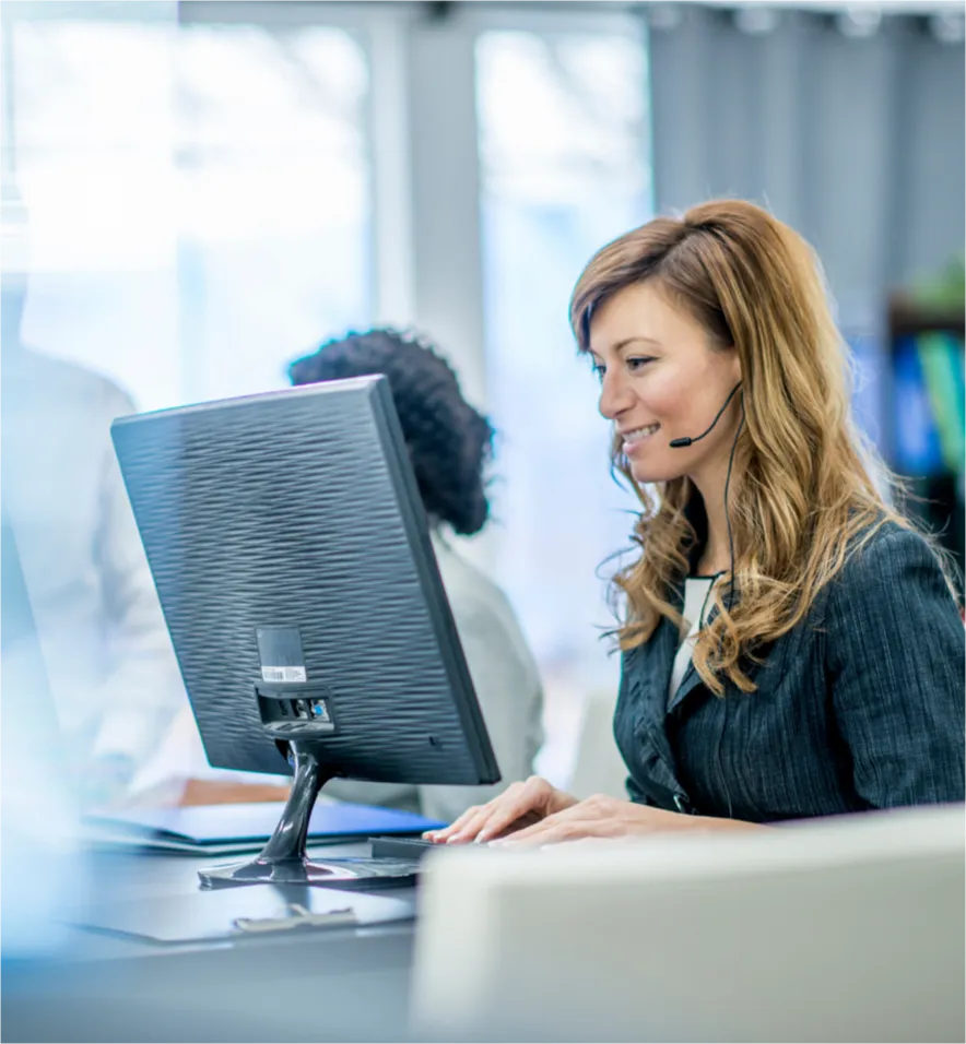 Woman wearing a headset smiles while working at a computer in a modern office setting.