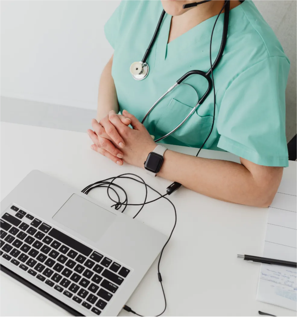 A healthcare professional in scrubs sits at a desk with a laptop, stethoscope, and folded hands.