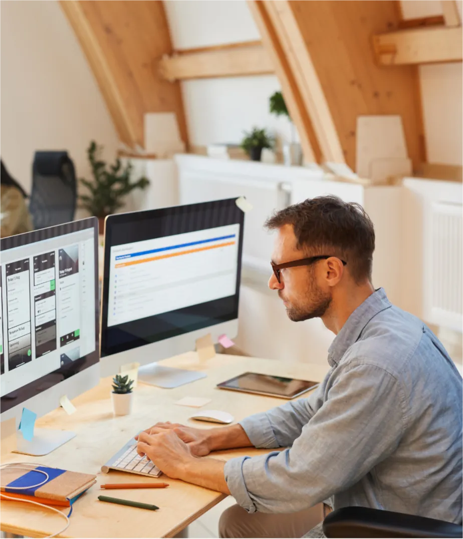 Man working at a desk with two monitors displaying websites in a bright, modern office with wooden beams.