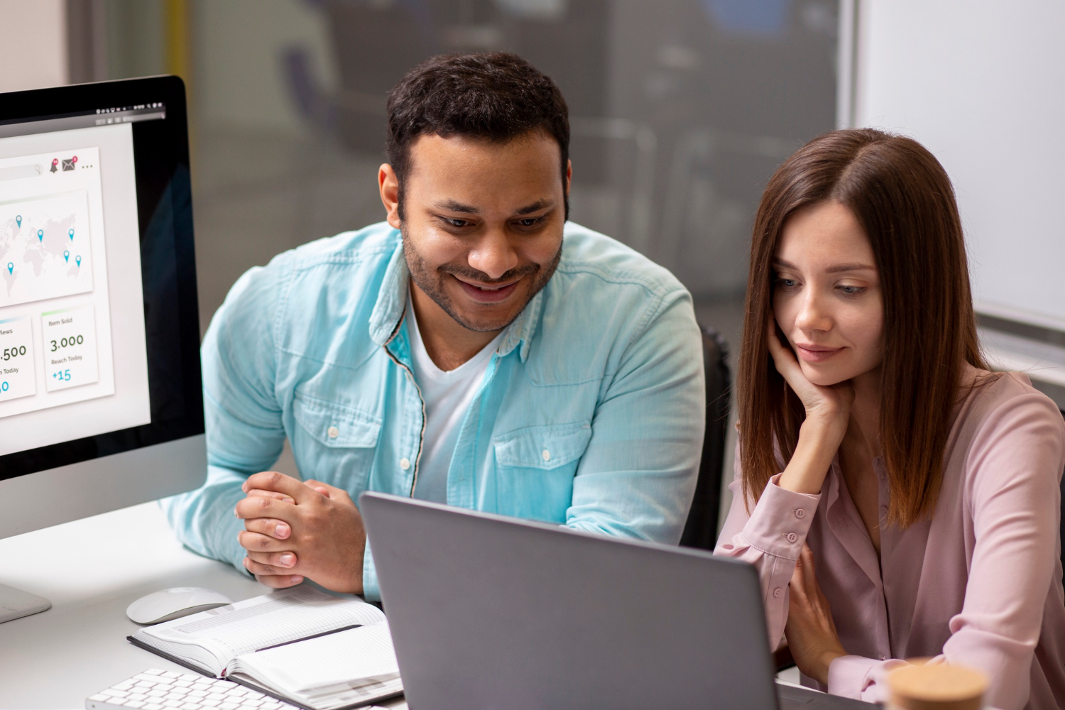 Two people sit at a desk, smiling and looking at a laptop, with a computer monitor beside them.