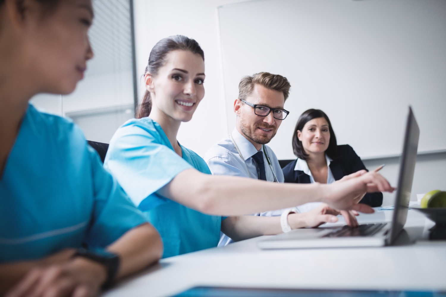 Three healthcare professionals and a businessperson smiling and working together at a laptop in a meeting room.