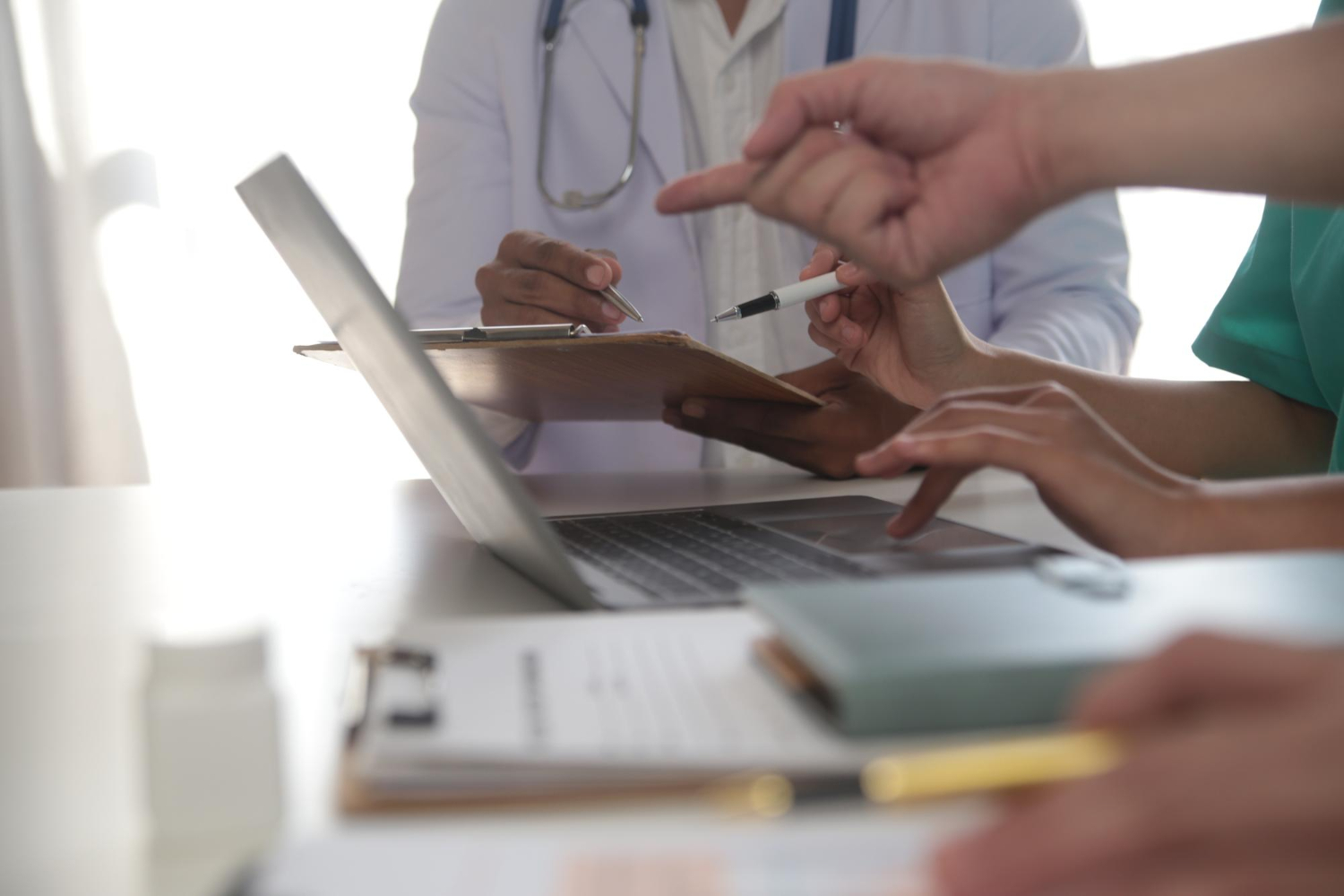 Doctors discussing medical notes with a clipboard and laptop, hands visible, in a bright office setting.