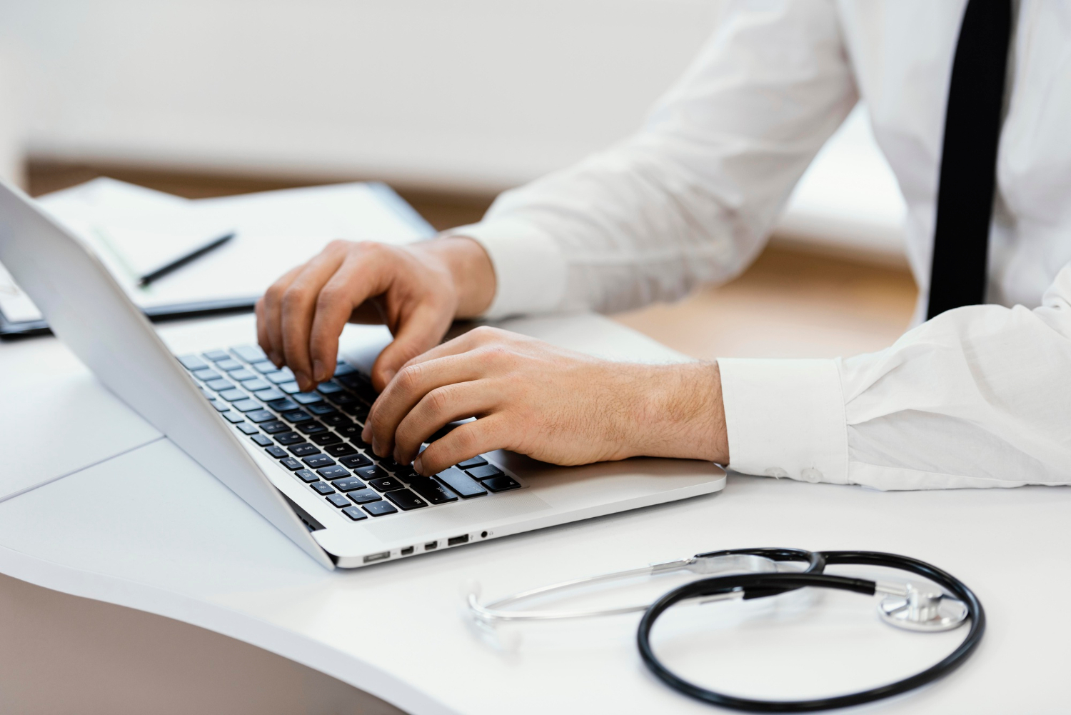 Person in a white shirt typing on a laptop at a desk with a stethoscope beside them.