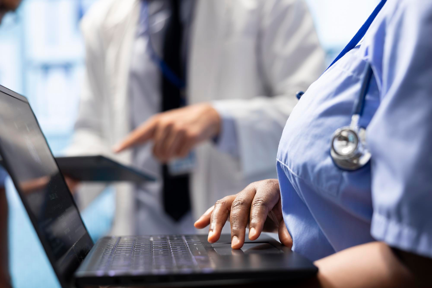 Close-up of a healthcare worker typing on a laptop with another doctor pointing at a tablet in the background.