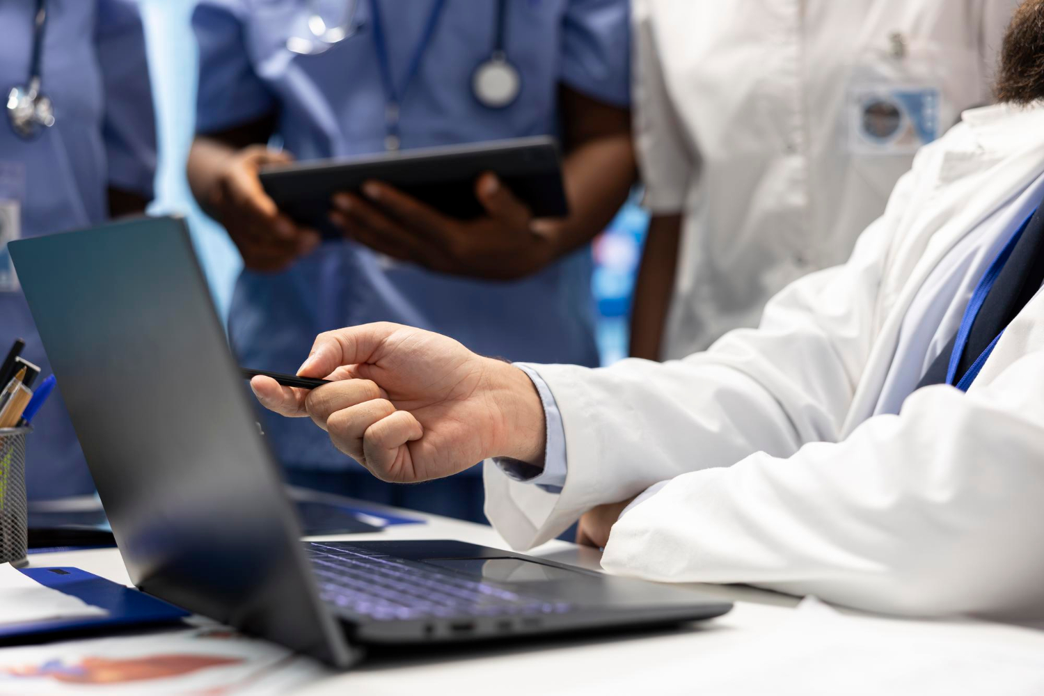 113893 A doctor points at a laptop screen while medical staff stand in the background with a clipboard.