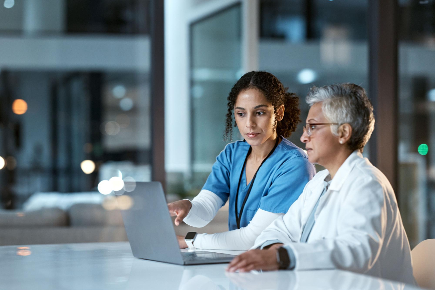 130872 Two female doctors in scrubs and lab coat discuss something while looking at a laptop in an office.